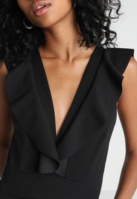 Svart topp med djup V-ringning, volangdetaljer vid axlarna och en slät tygstruktur. Närbilden betonar design och form.