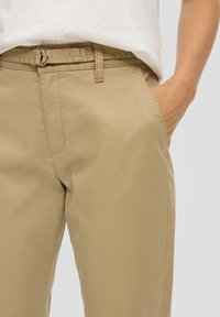 Persoon in beige broek met een stoffen riem, met de hand in de rechter voorzak, gecombineerd met een wit shirt.