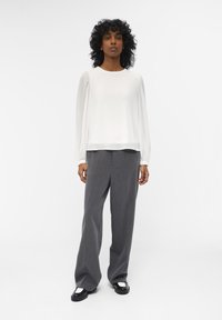 Blusa blanca de manga larga con puños fruncidos, combinada con pantalones de pierna ancha grises con un sutil patrón a rayas; zapatos negros con detalles blancos.