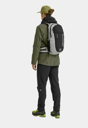 Rückansicht eines Mannes in einer grünen Jacke und schwarzen Hosen mit einem grau-schwarzen Ortovox-Rucksack, der ein Logo und verstellbare Schulterriemen aufweist.