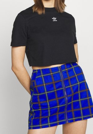 Kvinde iført en sort Adidas cropped t-shirt og en blåternet miniskørt med en lynlås foran, poserende mod en ensfarvet baggrund.