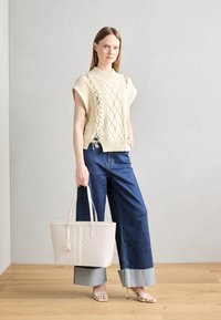 Pull en maille crème avec motif torsadé, manches larges ; associé à un jean large bleu foncé avec ourlet effiloché. Sac à main clair au design géométrique.