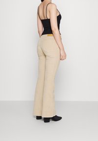 Beige utsvängda byxor med en texturerad yta, midjehög midja och två bakfickor. Modellen bär en svart tanktop och svarta ankelskoboots.