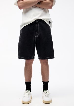 Person trägt schwarze Jeansshorts, weiße Sneakers mit beigen Sohlen, schwarze Socken und ein weißes T-Shirt, die Arme über der Taille verschränkt.