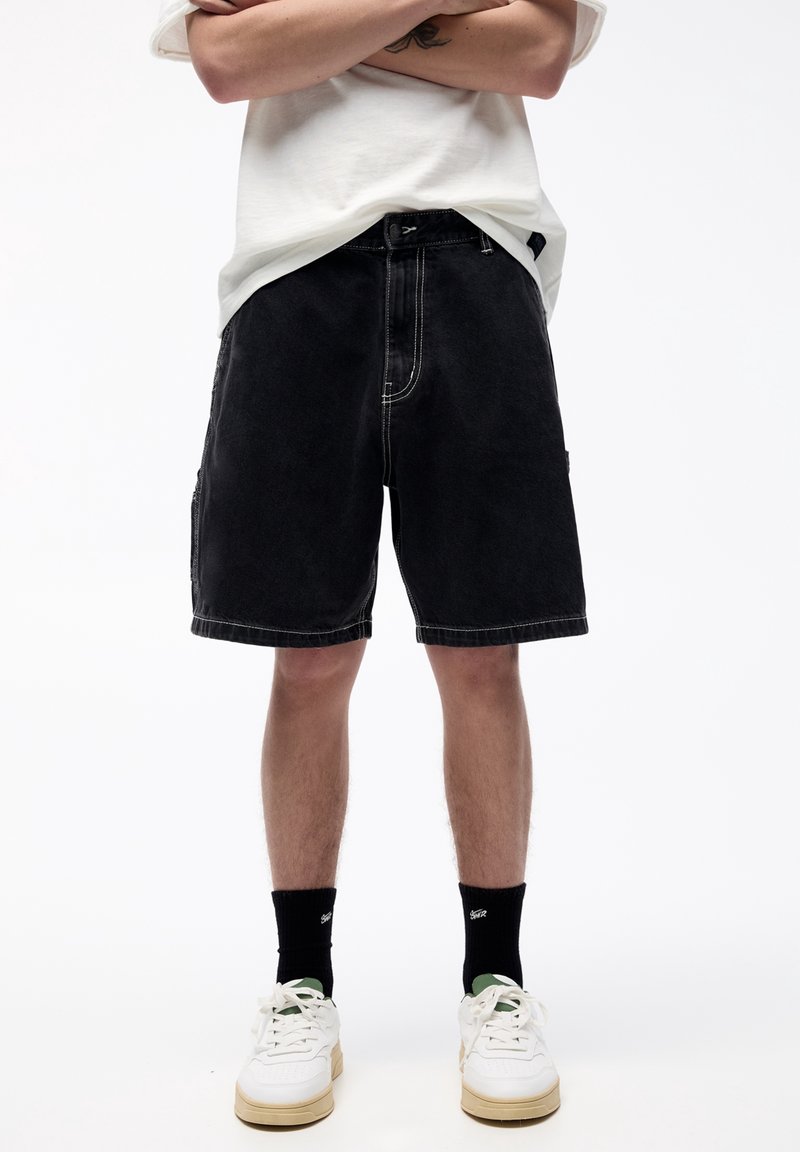 Personne portant un short en jean noir, des baskets blanches avec des semelles beige, des chaussettes noires et un T-shirt blanc, les bras croisés au-dessus de la taille.
