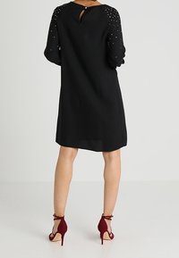 Robe noire avec des manches semi-transparentes et ornées, et un dos en clef de sol, associée à des talons bourgogne à lanières. Texture lisse dans l'ensemble.