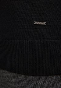 Schwarzer Strickpullover mit einem gerippten Saum und einem silbernen Metallschild mit der Aufschrift "JOOP!" am unteren Rand. Strukturierter Stoff mit feinen Musterdetails.