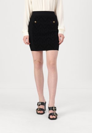 Woman wearing a black knee-length skirt with gold buttons, cream long-sleeve blouse, and black heeled sandals with large buckles.