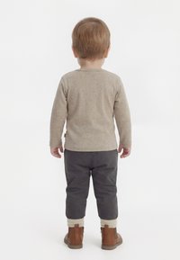 Batole stojící zády, oblečené v béžovém tričku s dlouhým rukávem, tmavých kalhotách, krémových ponožkách a hnědých kotníkových botách na bílém pozadí.