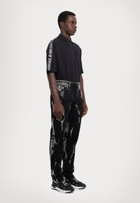 Melns īsām piedurknēm polo krekls ar rāvējslēdzēja apkakli un baltu logo uz piedurknes, apvienots ar melnām džinsām ar nolietojuma rakstu un melnām sporta kurpēm.