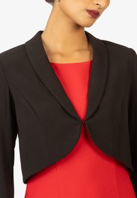 Blazer nero corto con colletto arrotondato, texture liscia e dettaglio di chiusura frontale, indossato sopra un abito rosso vivace.