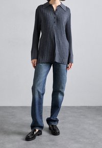 Person in einem dunkelgrauen, gerippten Langarmshirt, blauen, geraden Jeans und schwarzen, glänzenden Slippern steht auf einem grauen Boden vor einer hellgrauen Wand.