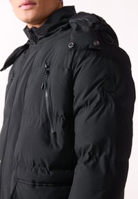 Chaleco acolchado negro con un cuello alto, bolsillos con cremallera y un parche de logo en la manga. Textura suave con un ajuste elegante.