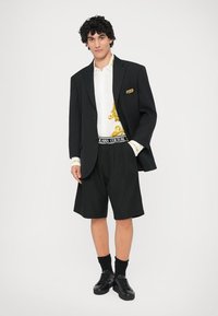 Črn blazer preko bele srajce z zlatim vzorcem, črne kratke hlače in temni čevlji. Vsebuje blagovno znamko na pasu in subtilne podrobnosti.