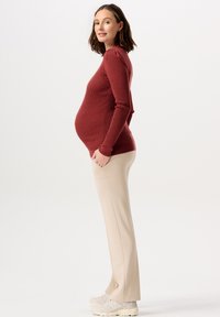 Graviditetsstickad tröja i mörk rostfärg, åtsittande silhuett. Parat med beige byxor och ljusa sneakers. Sidoprofilvy.