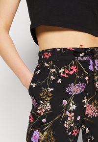 Černé květované kalhoty s páskem na stažení, s barevnými květinovými vzory v růžové, fialové a bílé na tmavém pozadí.