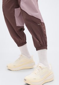 Καφέ και ροζ αθλητικό παντελόνι με ελαστικές μανσέτες, συνδυασμένο με ανοιχτοκίτρινα αθλητικά παπούτσια με υφασμένο άνω μέρος και λευκό λογότυπο.