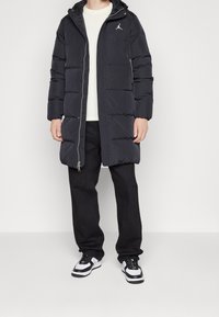 Zwarte pufferjas met een gewatteerd ontwerp, ritssluiting en capuchon. Witte logo op de linkerborst, gedragen over een licht shirt en zwarte broek. Sneakers zichtbaar.