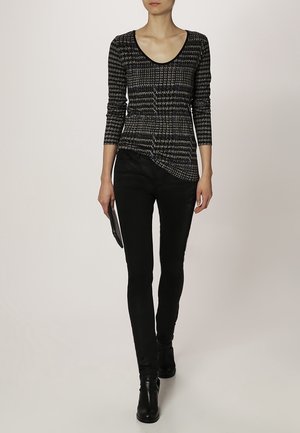 Mujer vestida con una blusa de manga larga negra y gris con patrón, pantalones ajustados negros, botas de tobillo negras, sosteniendo un pequeño bolso de mano negro.