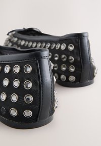 Chaussures noires sans lacets avec une tige texturée, ornées de clous argentés et de strass ronds réfléchissants pour plus de détails.