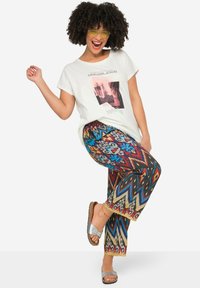 Biały t-shirt z grafiką w kwadratowym wzorze i napisem; kolorowe szerokie spodnie w wzory z niebieskimi, czerwonymi i żółtymi akcentami; srebrne klapki.