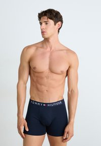 Homem sem camisa, com cabelo curto e encaracolado, vestindo cuecas boxer Tommy Hilfiger azul-marinho, em pé e olhando para a esquerda contra um fundo uniforme.