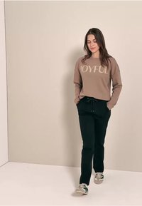 Hnědá mikina se slovem "JOYFUL", v kombinaci s černými tepláky. Model má na sobě šedé tenisky se zelenými akcenty. Hladké, pohodlné textury látek.