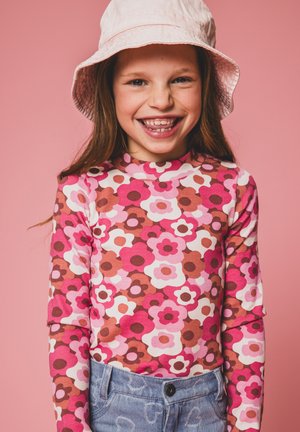 Langarmtop met een levendig bloemenpatroon in roze, bordeauxrood en wit. Gecombineerd met lichtblauwe jeans en een lichtroze bucket hat.