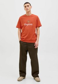 Orange bomulds t-shirt med rund halsudskæring og korte ærmer, prydet med en hvid grafisk tekst "Originals" og solstråledesign. Båret sammen med brune bukser.