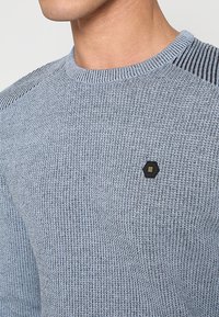 Gebreide trui in lichtblauw met textuurstof, voorzien van een geribde halslijn en een zeshoekig merkembleem in zwart op de borst.