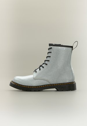 Lichtgrijze leren veterboot met zwarte zool, gele stiksels en trek lus, in profiel weergegeven op een neutrale achtergrond.