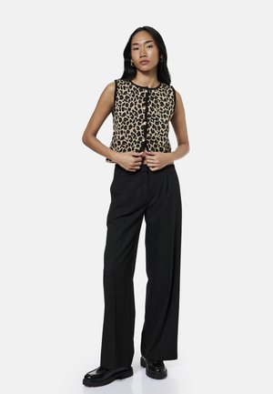 Vrouw staand, gekleed in een mouwloze button-down top met luipaardprint, zwarte brede broekspijpen en zwarte stevige schoenen tegen een effen achtergrond.