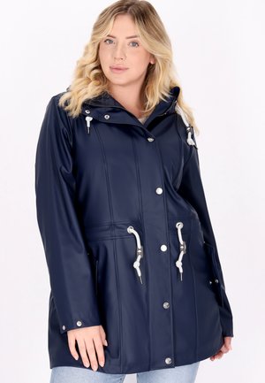Vrouw met blond haar die een marineblauwe regenjas draagt met witte trekkoorden en zilveren knopen tegen een effen witte achtergrond.