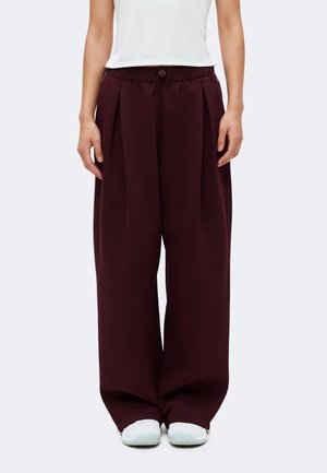 Personne portant un pantalon bordeaux à jambes larges avec une taille à cordon et des baskets blanches, montrée de la taille aux pieds sur un fond uni.