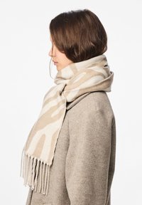 Beige tørklæde med et lyst brunt abstrakt mønster, lavet af blødt tekstil, med frynser i enderne, båret over en grå frakke, hængende løst.