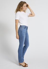 Naine seisab külgvaates, kandes valget lühikese varrukaga särki, siniseid teksaseid ja pruune avatud varbaga kontsakingi, taustaks sirge ja lihtne taust.