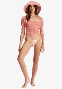 Top curto floral vermelho com mangas bufantes, combinado com um biquíni floral em rosa e amarelo. A modelo também está a usar um chapéu de abas largas combinado.