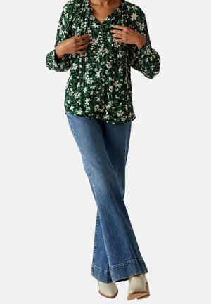 Frau in einer grünen Blumenbluse, blauen Schlagjeans und weißen, taillierten Absatzstiefeln, die mit übergeschlagenen Beinen steht.