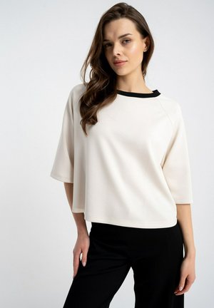 Kobieta ubrana w luźną białą bluzkę z czarnym kołnierzem i czarne spodnie, stojąca z jedną ręką opartą na biodrze na jednolitym tle.