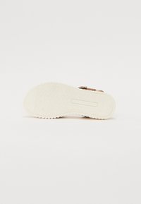 Bruine leren sandaal met een witte rubberen zool. De zool heeft een gestructureerd patroon en een glad deel. Verstelbare gespsluiting aan de zijkant.