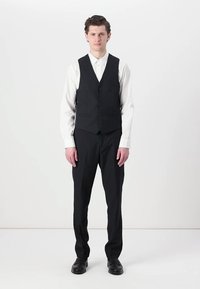 Gilet noir avec six boutons, porté sur une chemise blanche à manches longues, associé à un pantalon noir et des chaussures vernies. Design simple et ajusté.