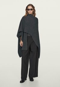 Žena v tmavém oversize ponču, širokých černých kalhotách, černých botách a obdélníkových slunečních brýlích, stojící na pozadí s jednoduchým světlým motivem.