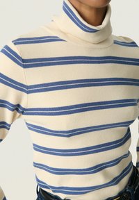 Jersey de cuello alto beige con rayas horizontales azules, usado metido dentro de unos jeans azul oscuro con un cinturón negro.