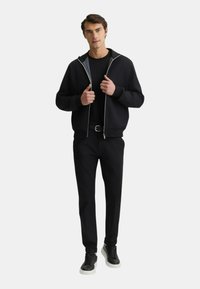 Giacca bomber nera con zip sopra una maglietta nera, abbinata a pantaloni neri e sneakers nere con suole bianche. Linee pulite e design semplice.