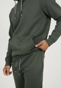 Sweat à capuche vert foncé avec poche kangourou, cordon de serrage ajustable avec accent à motif, assorti à un pantalon de survêtement fuselé coordonné.