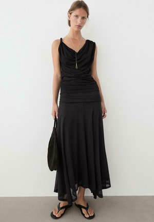 Kobieta nosząca bezrękawnikowy czarny drapowany top, długą czarną spódnicę, czarne sandały, trzymająca czarną torbę, stojąca przy białej ścianie.