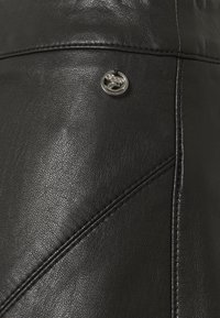 Lähikuva mustast nahkakudost, millel on detailsed õmblused ja väike ümar metallist logo, mis on kinnitatud õmbluse lähedale.