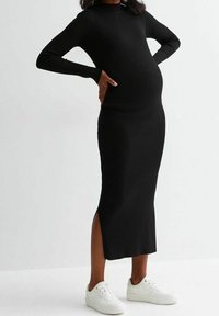 Gravid person iförd en svart ribbad långärmad midiklänning med sidoöppning och vita sneakers, med händerna vilande på ländryggen.