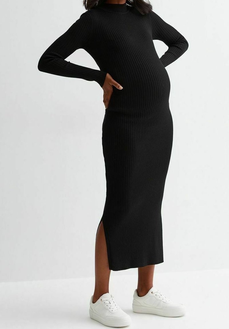 Gravid person iförd en svart ribbad långärmad midiklänning med sidoöppning och vita sneakers, med händerna vilande på ländryggen.