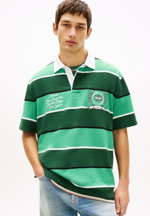 Jonge man met groen en donkergroen gestreept rugbyshirt met witte kraag en geborduurde stadsnamen en logo, gecombineerd met lichtblauwe jeans.